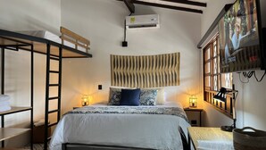 A compact hotel room with a bunk bed, a large bed with patterned bedding, a wall-mounted TV, and a small table with a basket.