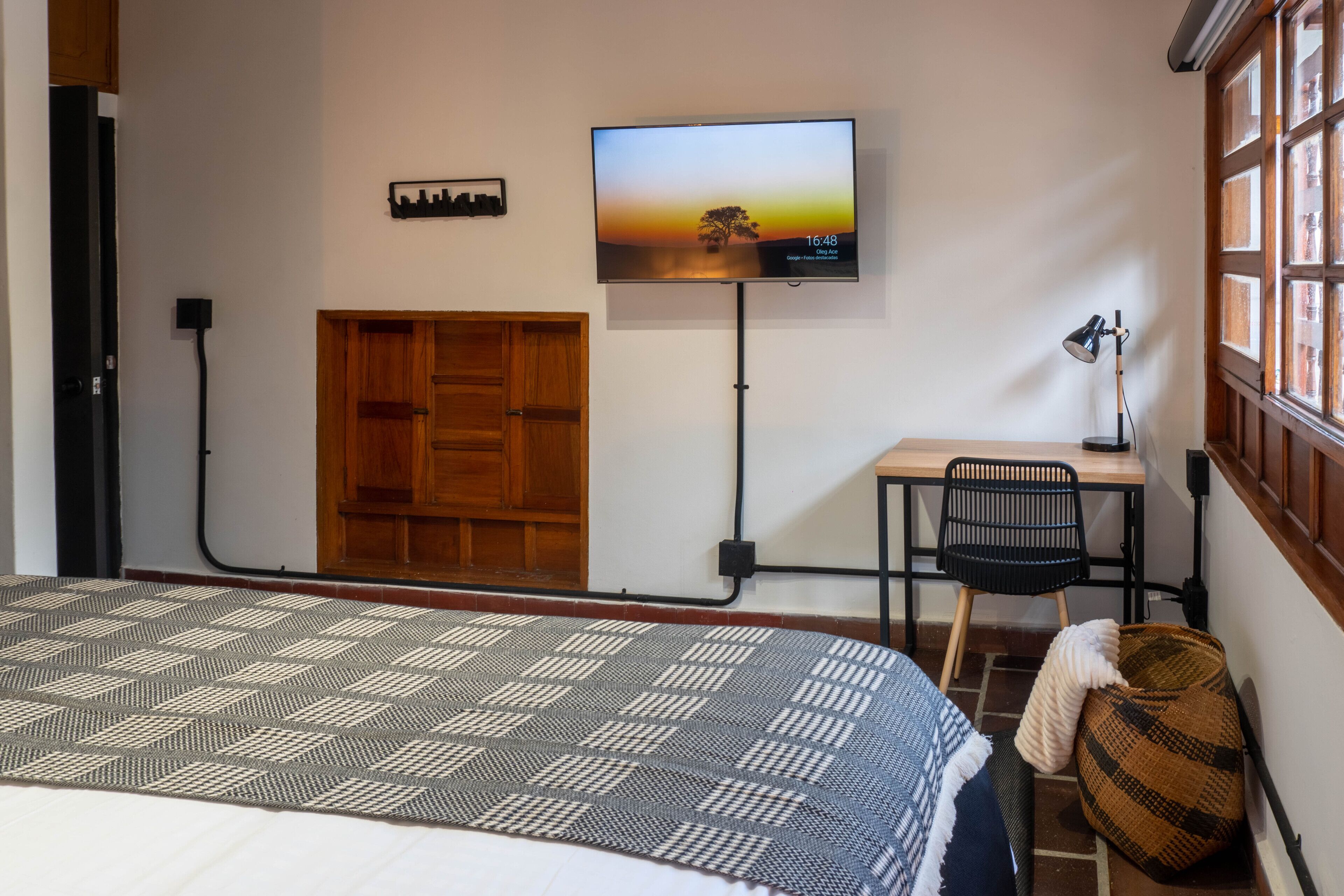 Una habitación con cama, escritorio, silla, televisión montada en la pared y ventana con marcos de madera.