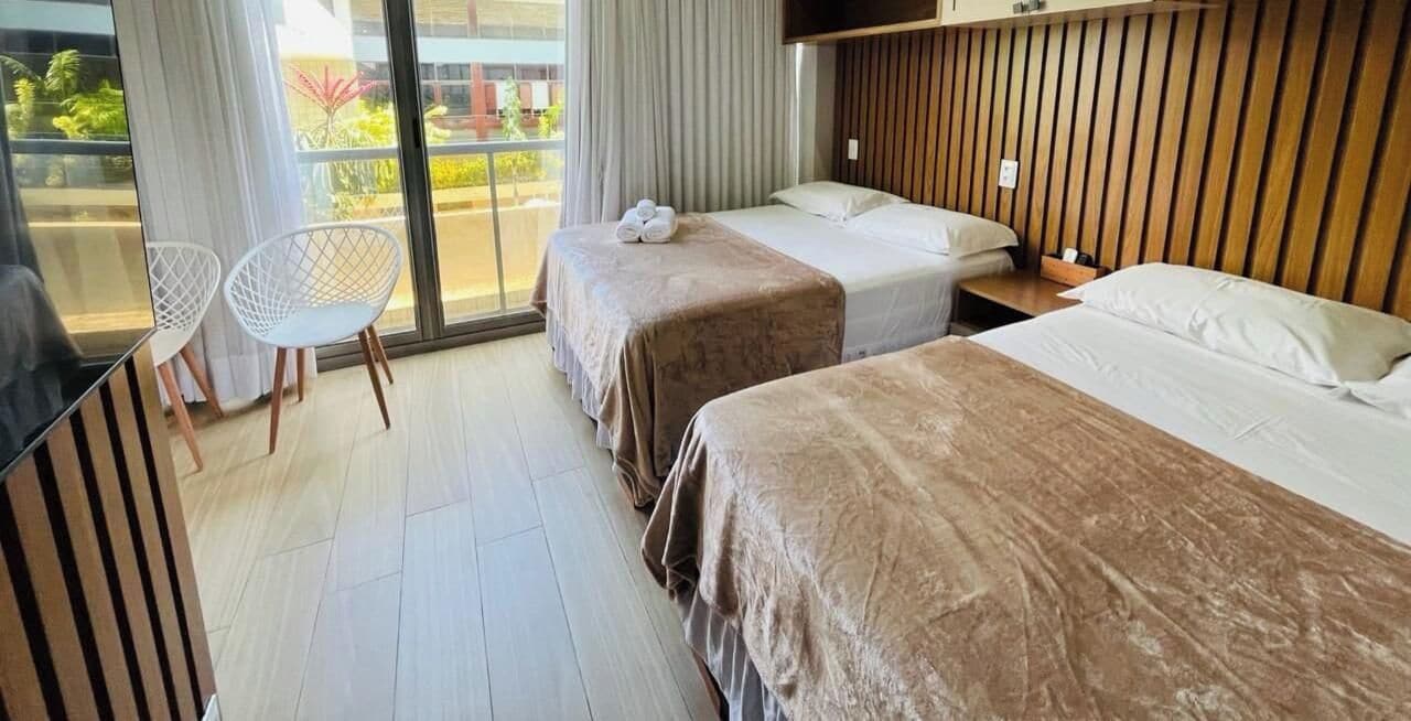 Habitación de hotel con dos camas, una zona de estar con un sillón y vistas al exterior.