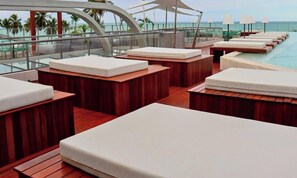 Zona junto a la piscina con tumbonas de madera y barandilla de cristal con vistas al mar.
