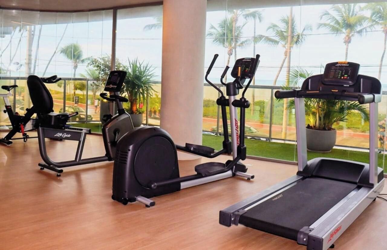 Un gimnasio con equipo cardiovascular, incluyendo cinta de correr, elíptica y bicicleta estática, con grandes ventanales que ofrecen vistas a palmeras.