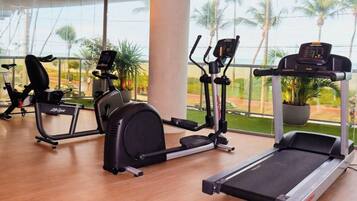 Un gimnasio con equipo cardiovascular, incluyendo cinta de correr, elíptica y bicicleta estática, con grandes ventanales que ofrecen vistas a palmeras.