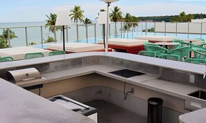 Zona junto a la piscina con un bar de hormigón, un cubo de basura y sillas de plástico verde.