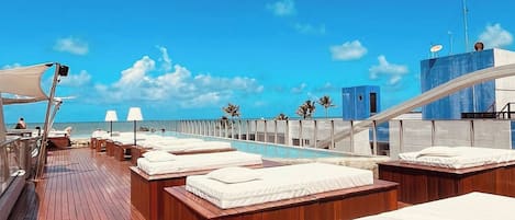 A sunlit deck with wooden flooring, white cushioned seating, and a clear view of the ocean.