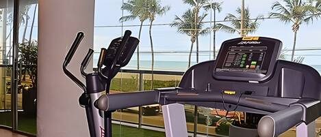 Una bicicleta estática con pantalla digital, ubicada cerca de un ventanal con vistas a una playa y palmeras.