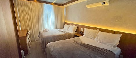 A hotel room with two beds, a ceiling air conditioner, and a window with curtains.