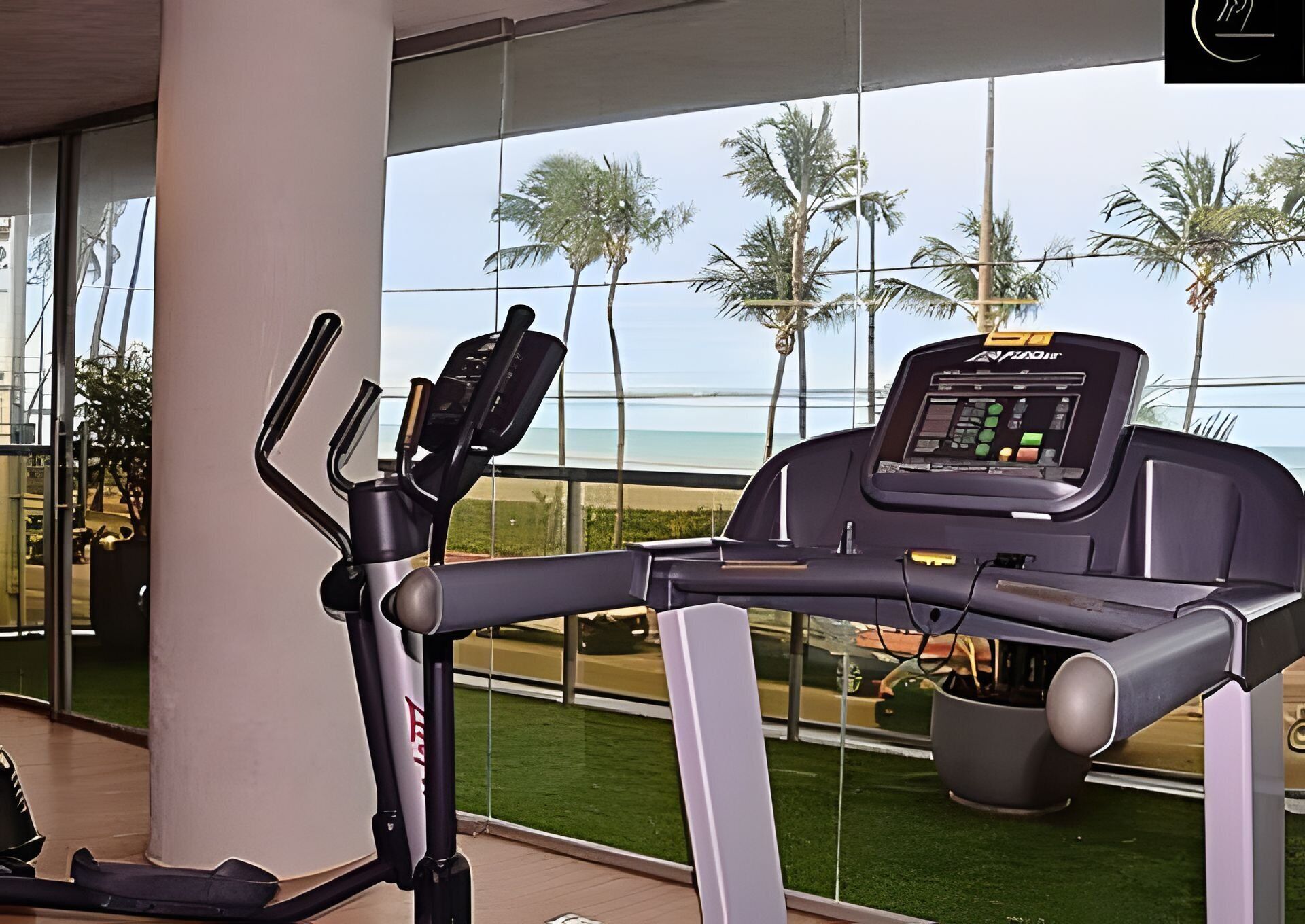 Una bicicleta estática con pantalla digital, ubicada cerca de un ventanal con vistas a una playa y palmeras.