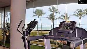 Un vélo d’appartement doté d’un écran numérique, placé près d’une grande fenêtre donnant sur une plage bordée de palmiers.