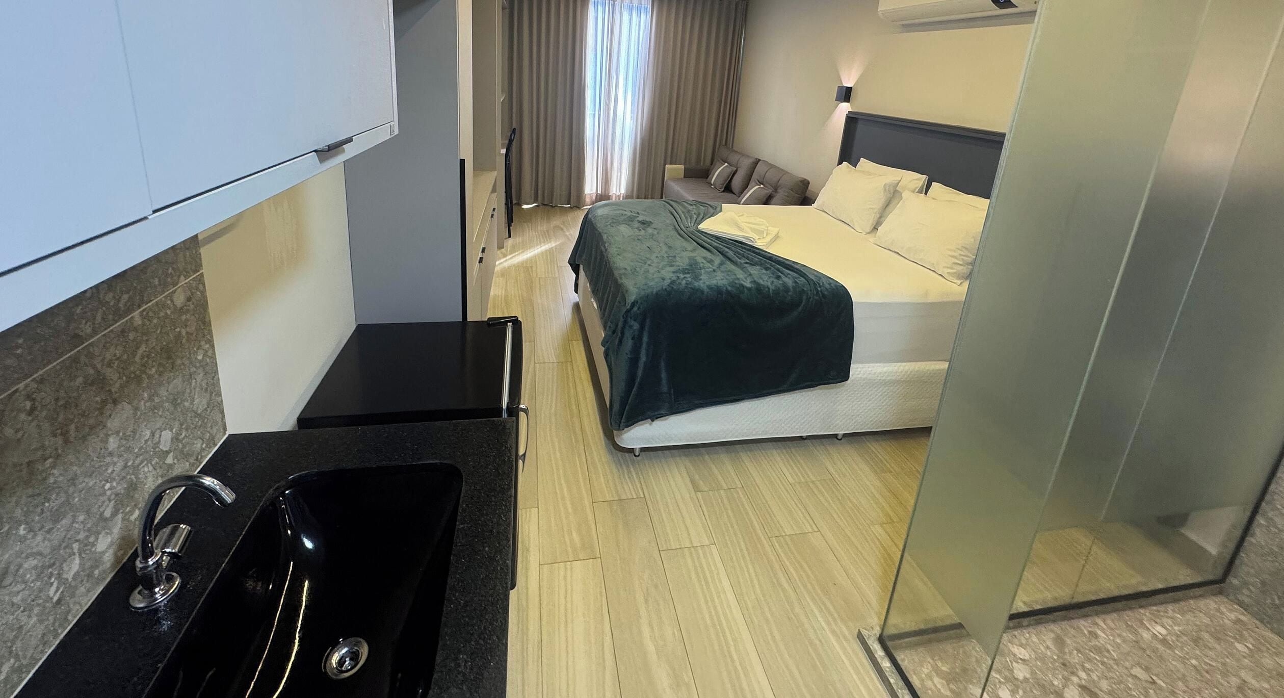 A compact hotel room with a kitchenette, a bed with a green bedspread, and a window with curtains.