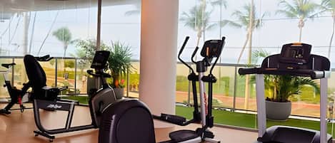 A gym with cardio machines, a treadmill, and large windows with a view of palm trees.