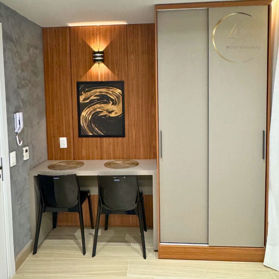 A room with a wooden wall panel, a framed artwork, a desk with two chairs, and a wardrobe with a circular logo.