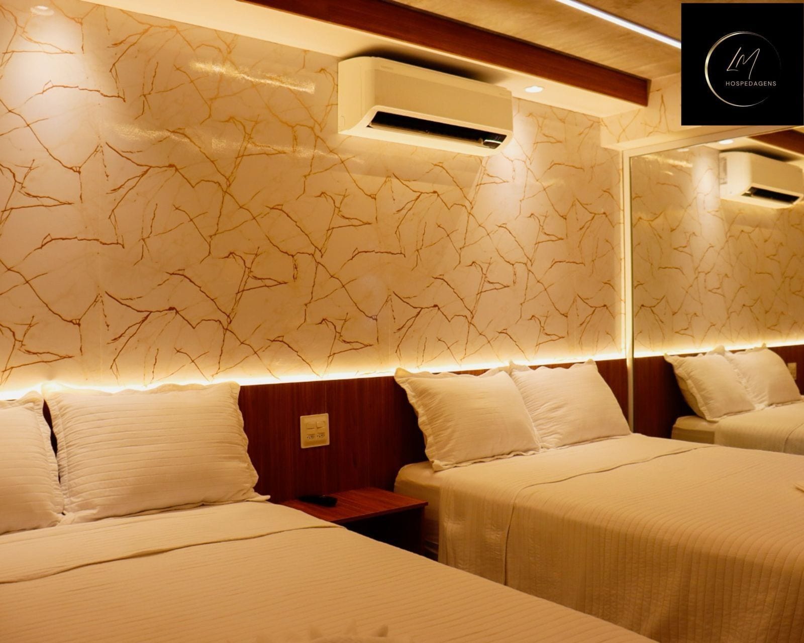 A hotel room with two beds, a wooden headboard, a wall-mounted air conditioning unit, and a bedside table.