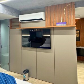 Un dormitorio con televisor empotrado, equipo de aire acondicionado, un cubo de basura y pared de paneles de madera.
