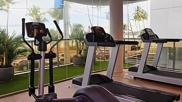 Ein Fitnessstudio mit Laufbändern und Trainingsgeräten, große Fenster mit Blick auf Palmen und einen Parkplatz.
