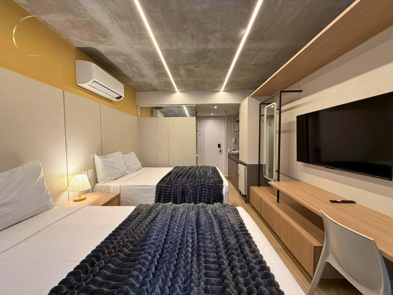 A modern hotel room with two beds, a TV, a desk, and a bathroom visible in the background.