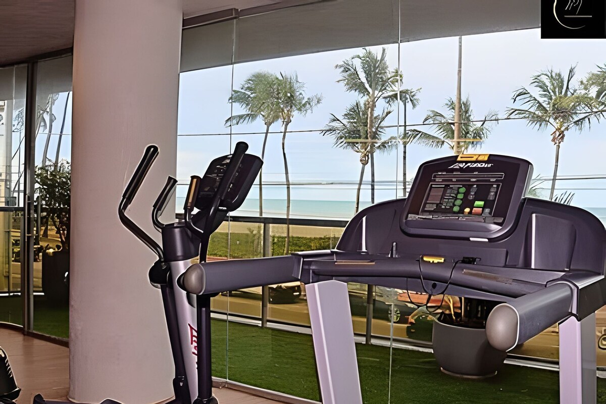 Uma bicicleta ergométrica com visor digital, posicionada perto de uma grande janela com vista para uma praia e palmeiras.