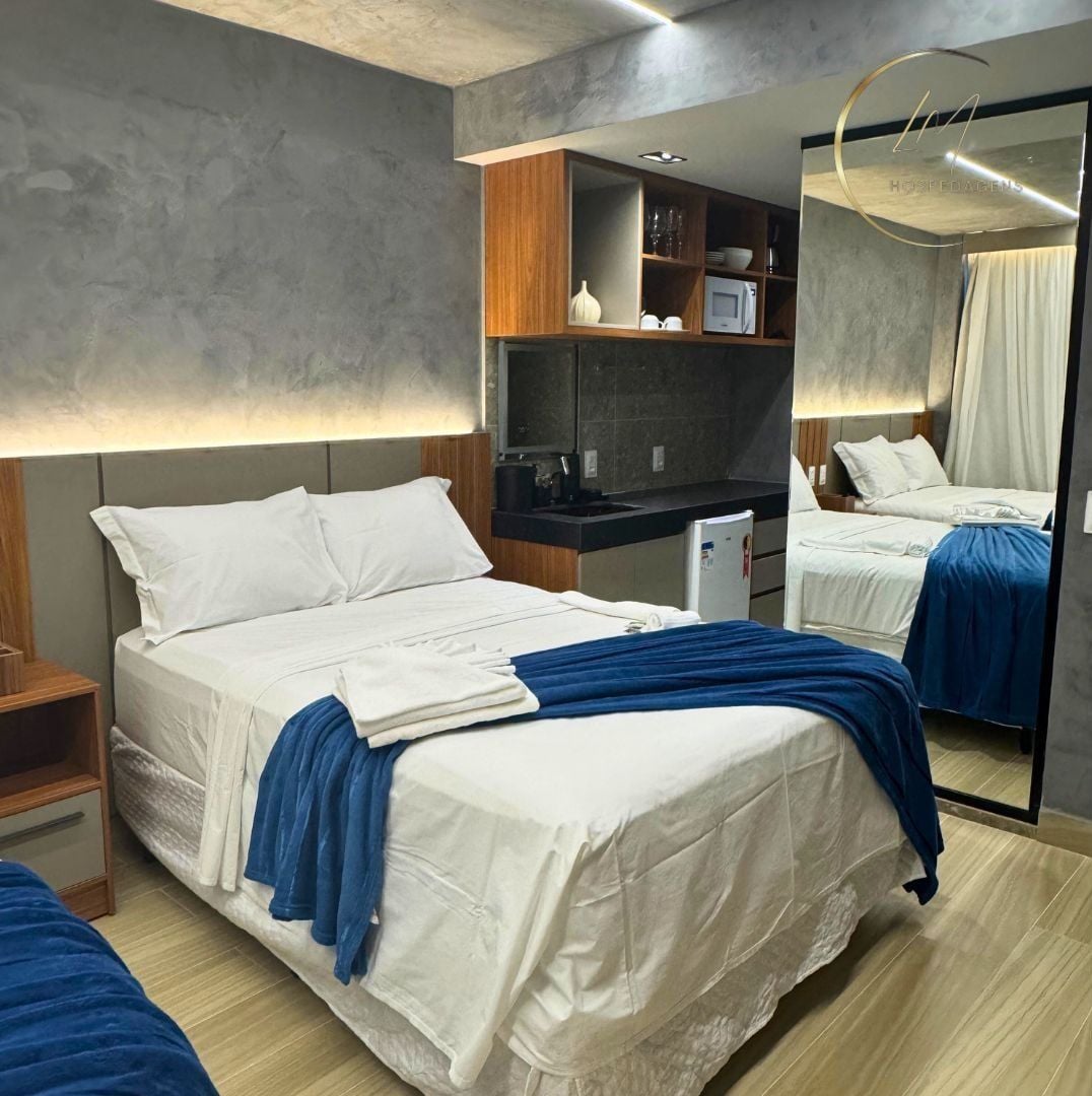 A modern hotel room with a large bed, a small kitchenette, a mirror, and a wooden headboard.