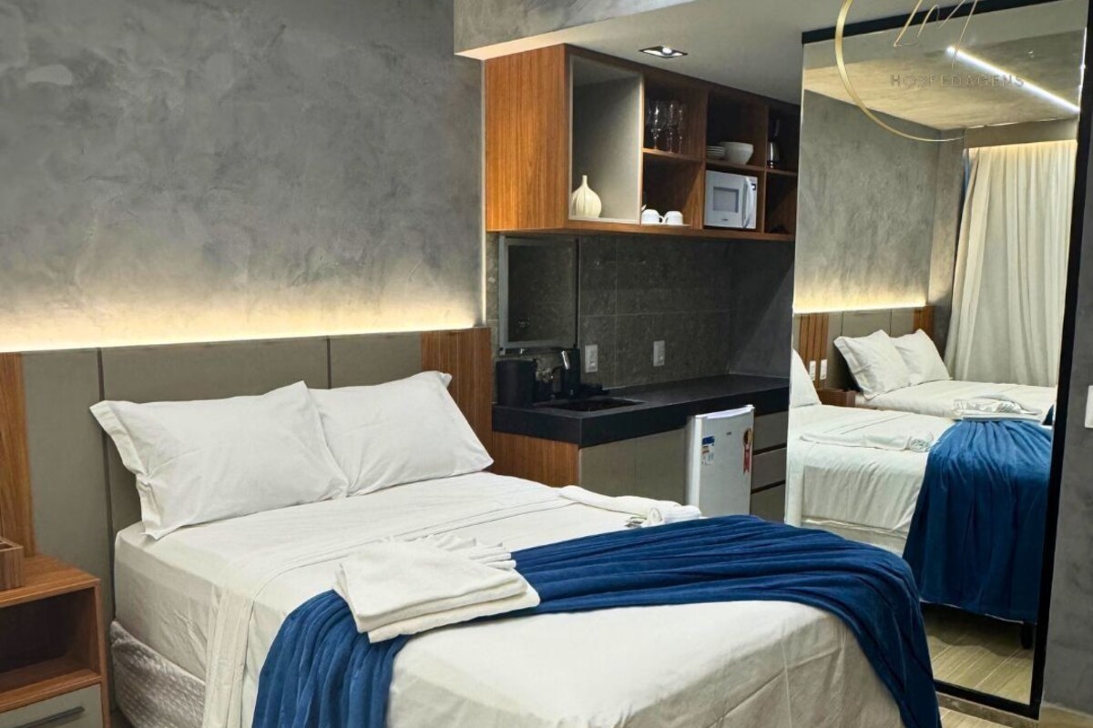 Quarto de hotel moderno com uma cama grande, uma kitchenette pequena, um espelho e uma cabeceira de madeira.