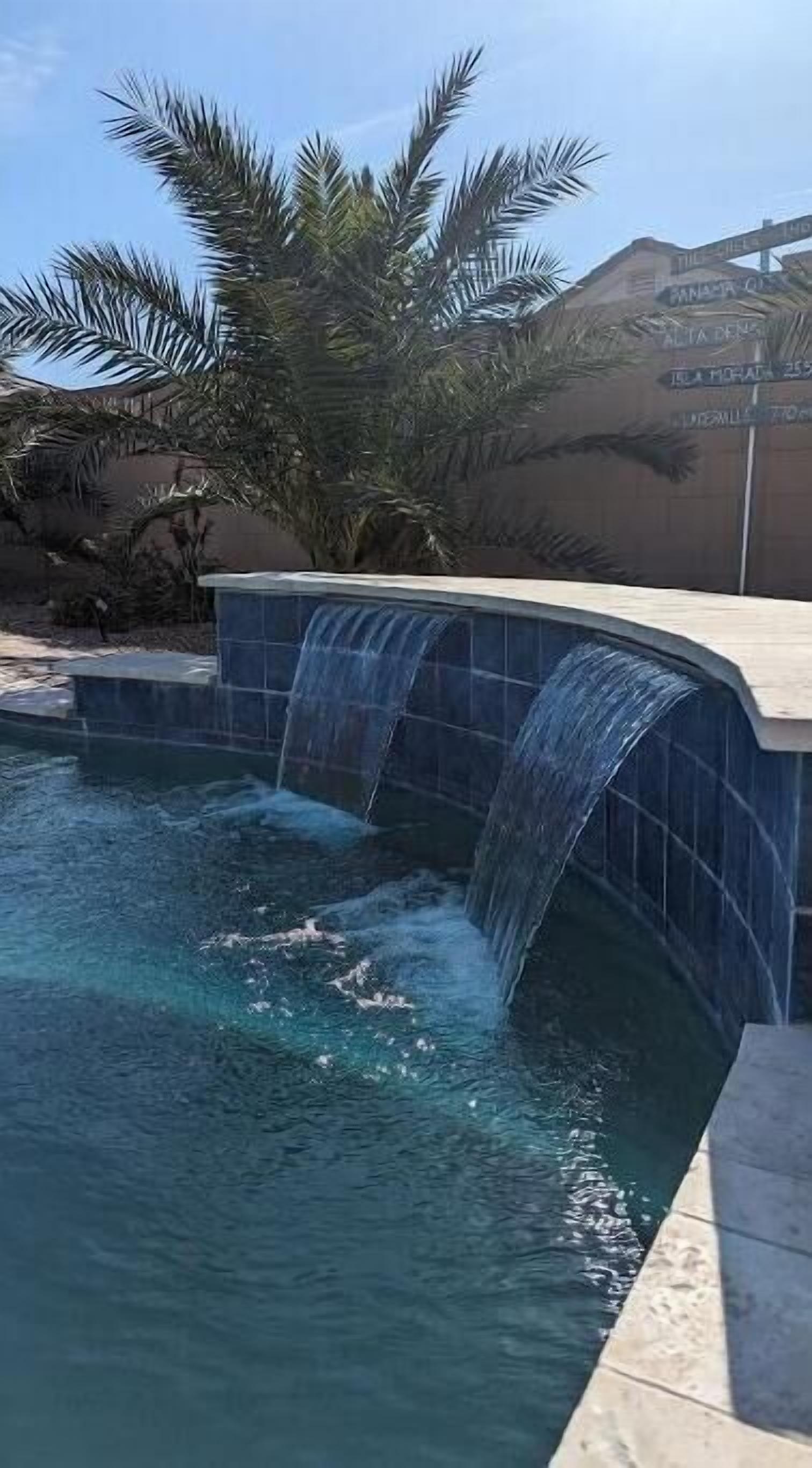 Ein Swimmingpool mit Wasserfall, umgeben von Palmen und einer Steinmauer.