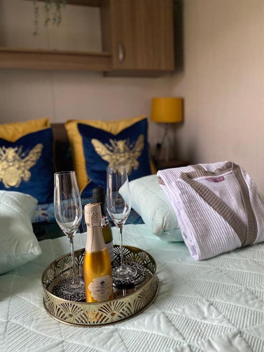 Een bed met een decoratieve dienblad waarop een fles en twee glazen staan, kussens met een familiewapen en een lamp op de achtergrond.