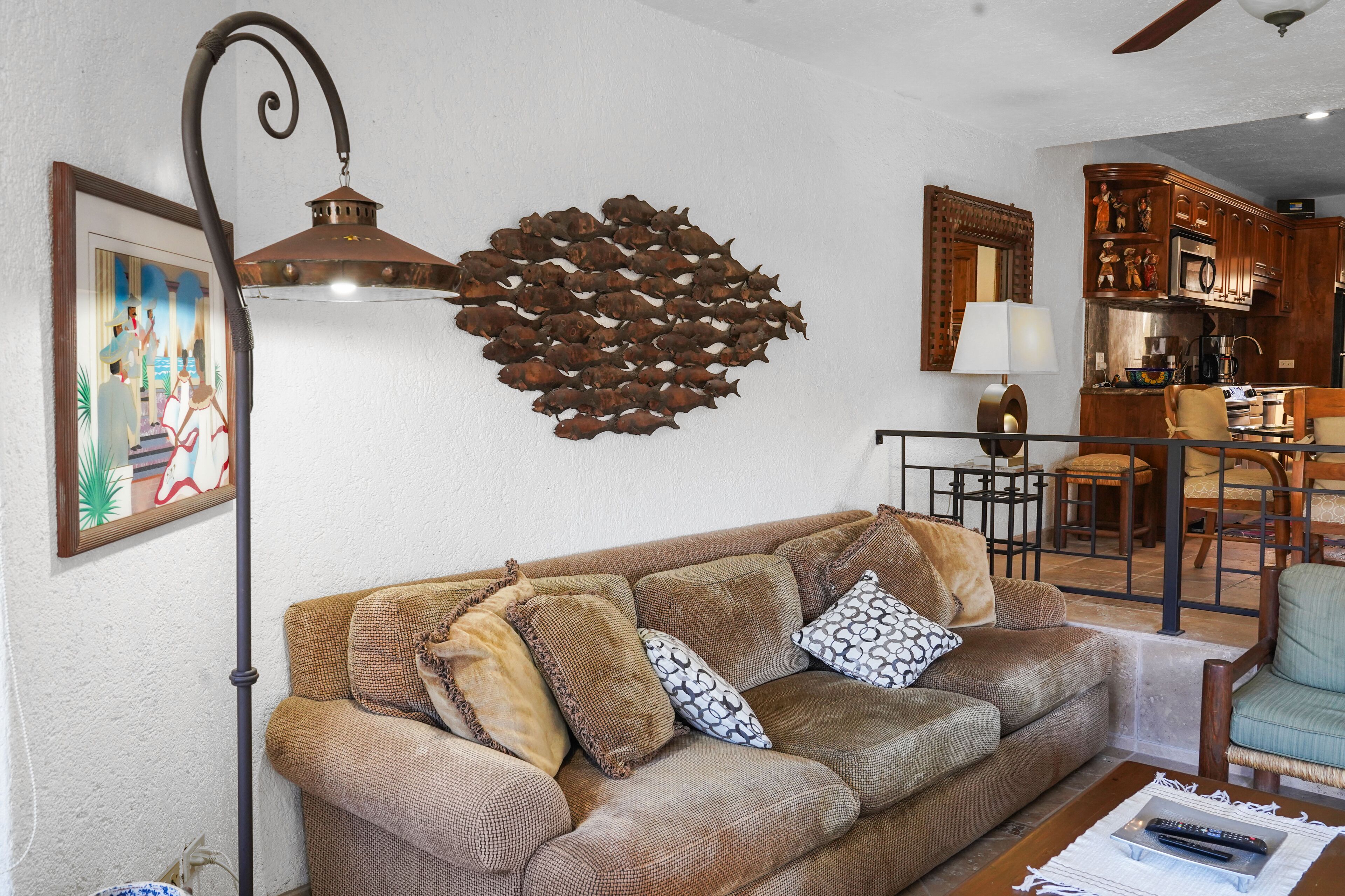 A living room with a brown sofa, patterned cushions, a wall art piece, and a kitchen area in the background.
