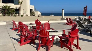 A patio with red Adirondack chairs and a fire pit, overlooking a beach with a clear sky. - 2 bedroom condo, marina view in Terrasol Beach Resort, 15 min walk to everything (Cabo San Lucas)