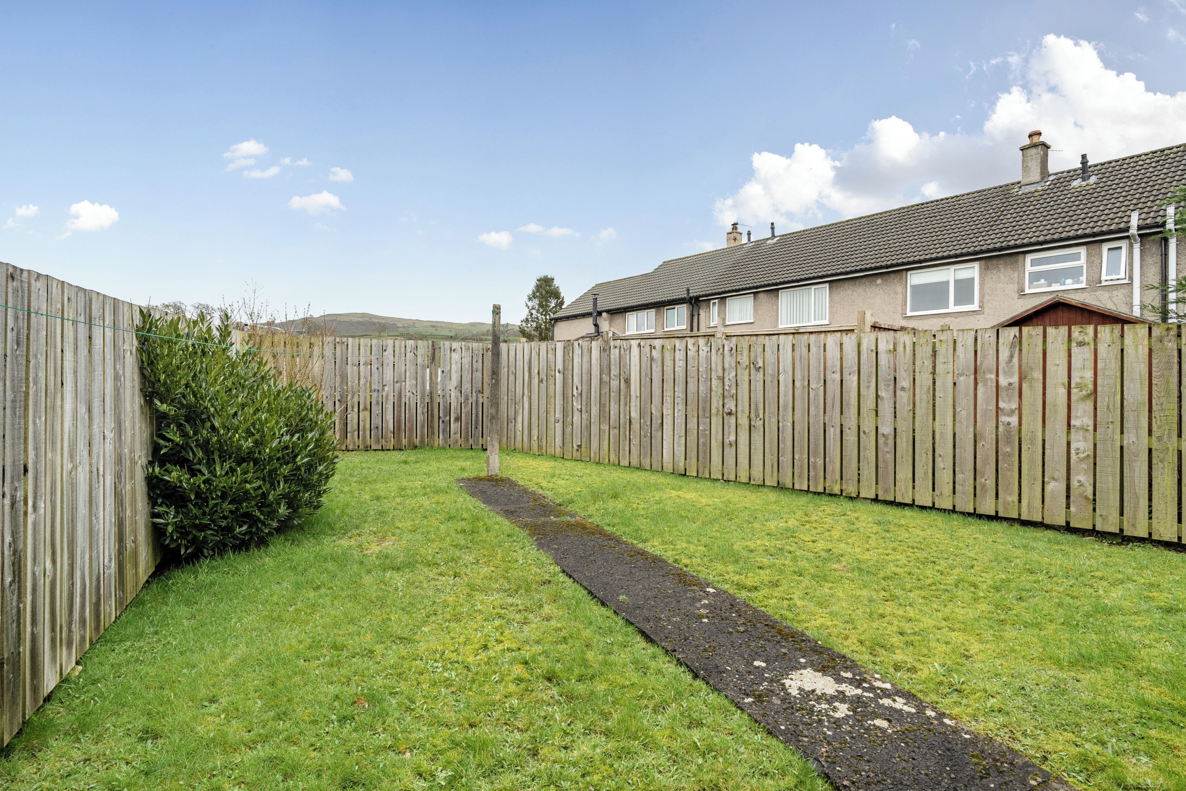Un jardin à l’arrière d’une maison, avec une clôture en bois, un chemin dallé et une maison dont le toit est recouvert de tuiles.