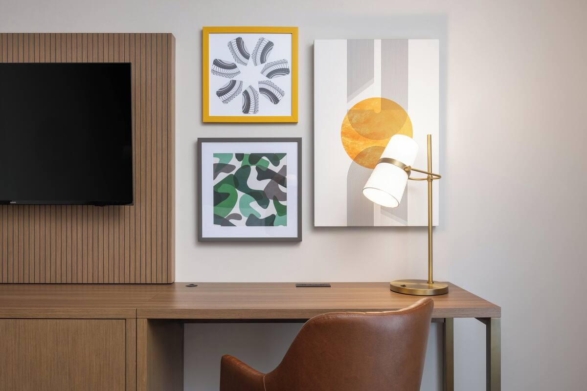 Une chambre moderne avec un bureau en bois, un fauteuil en cuir, un téléviseur à écran plat et trois œuvres d’art encadrées accrochées au mur.