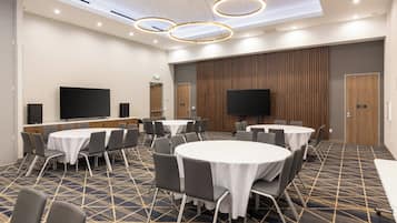 A conference room with round tables, chairs, a large flat-screen TV, and a patterned carpet.
