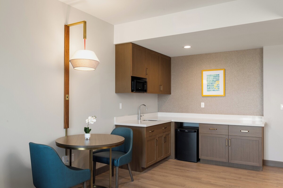 A modern kitchen with wooden cabinets, a black microwave, and a sink. There is a dining table with two chairs and a small potted plant.