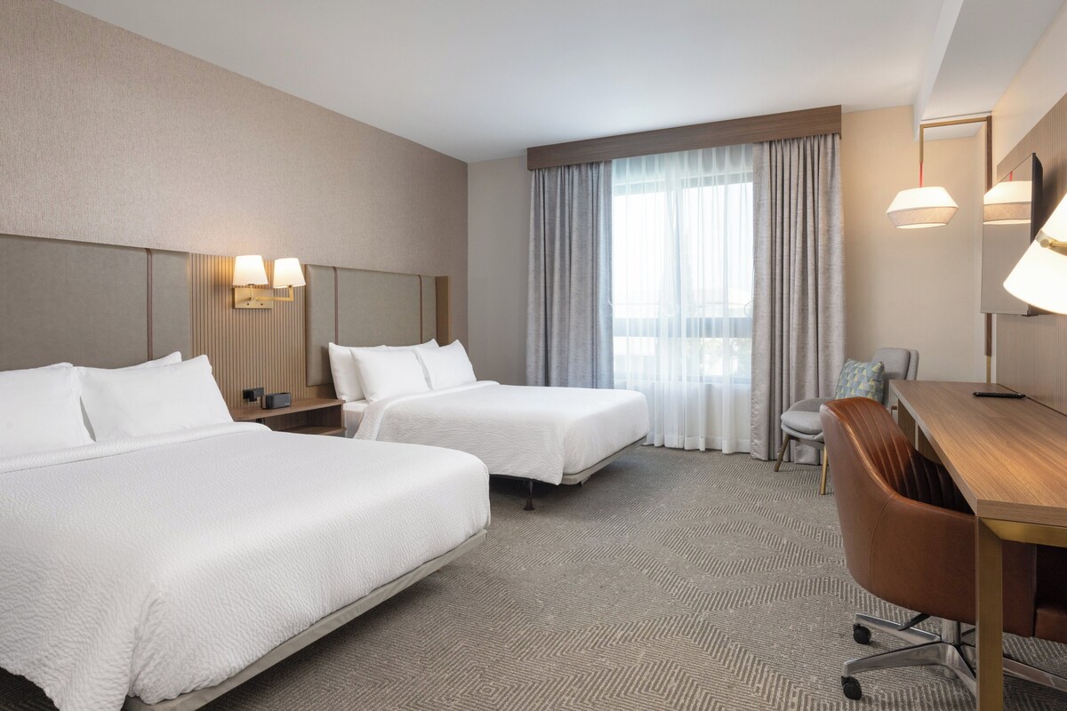 A hotel room with two beds, a desk, a chair, a nightstand, and a large window with curtains.