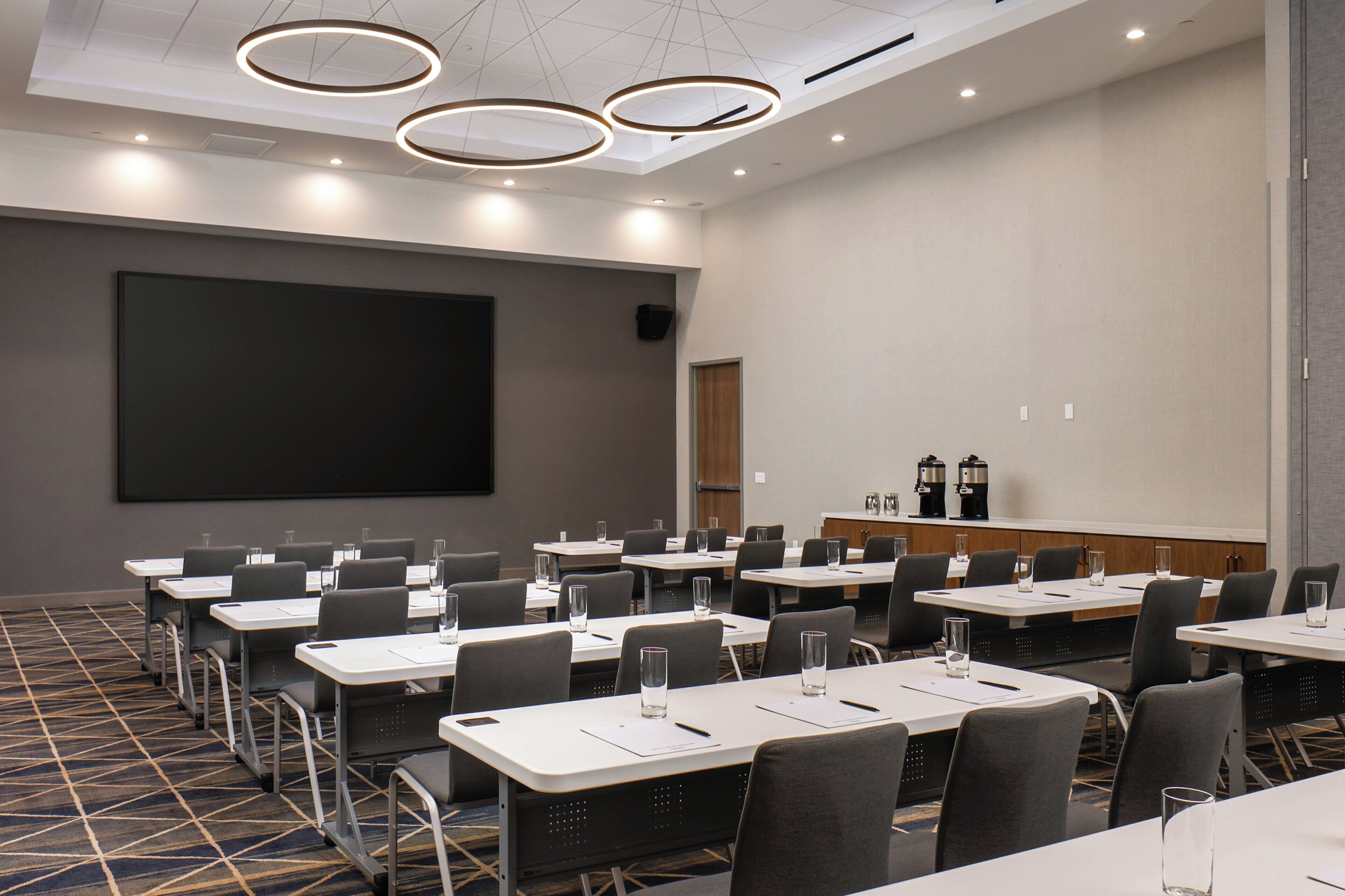A conference room with multiple tables, chairs, a large screen, and a coffee station.