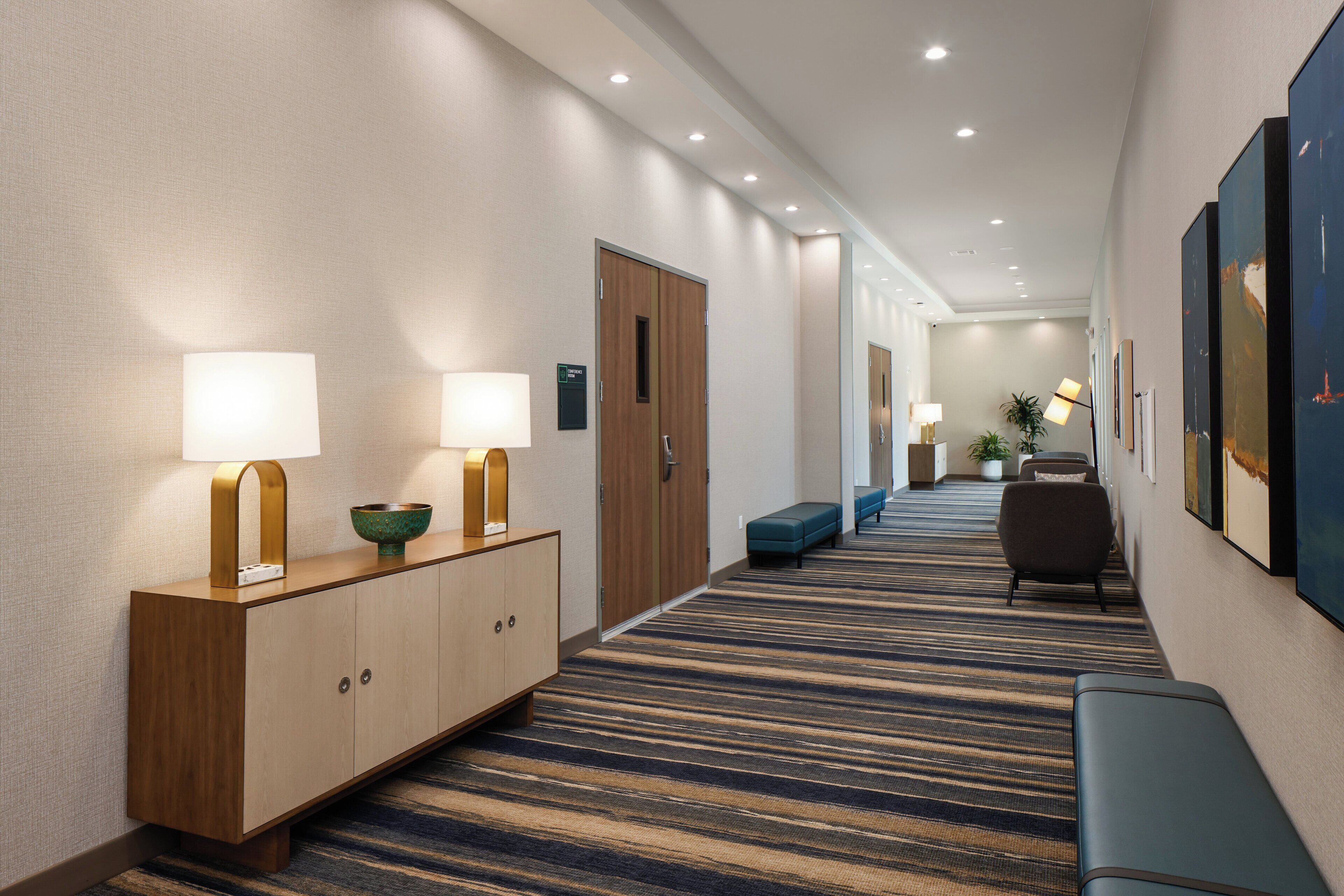 Un pasillo de hotel con suelo alfombrado, un banco de madera, una recepción con lámpara y cuadros en la pared.