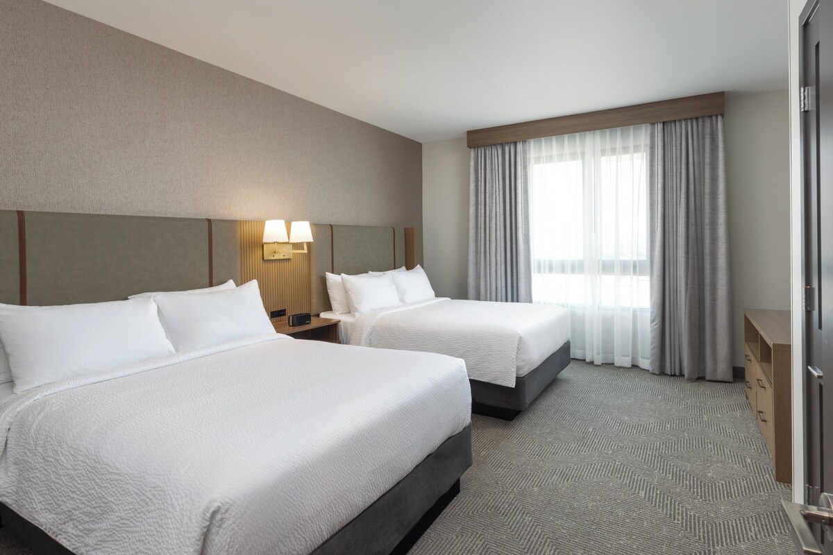 A hotel room with two beds, a nightstand with a lamp, a wooden dresser, and a large window with curtains.