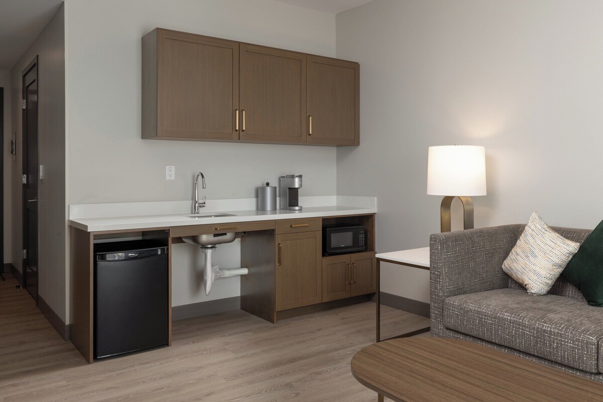 A compact kitchen with wooden cabinets, a dishwasher, a microwave, and a coffee maker. There is a sofa and a lamp in the living area.