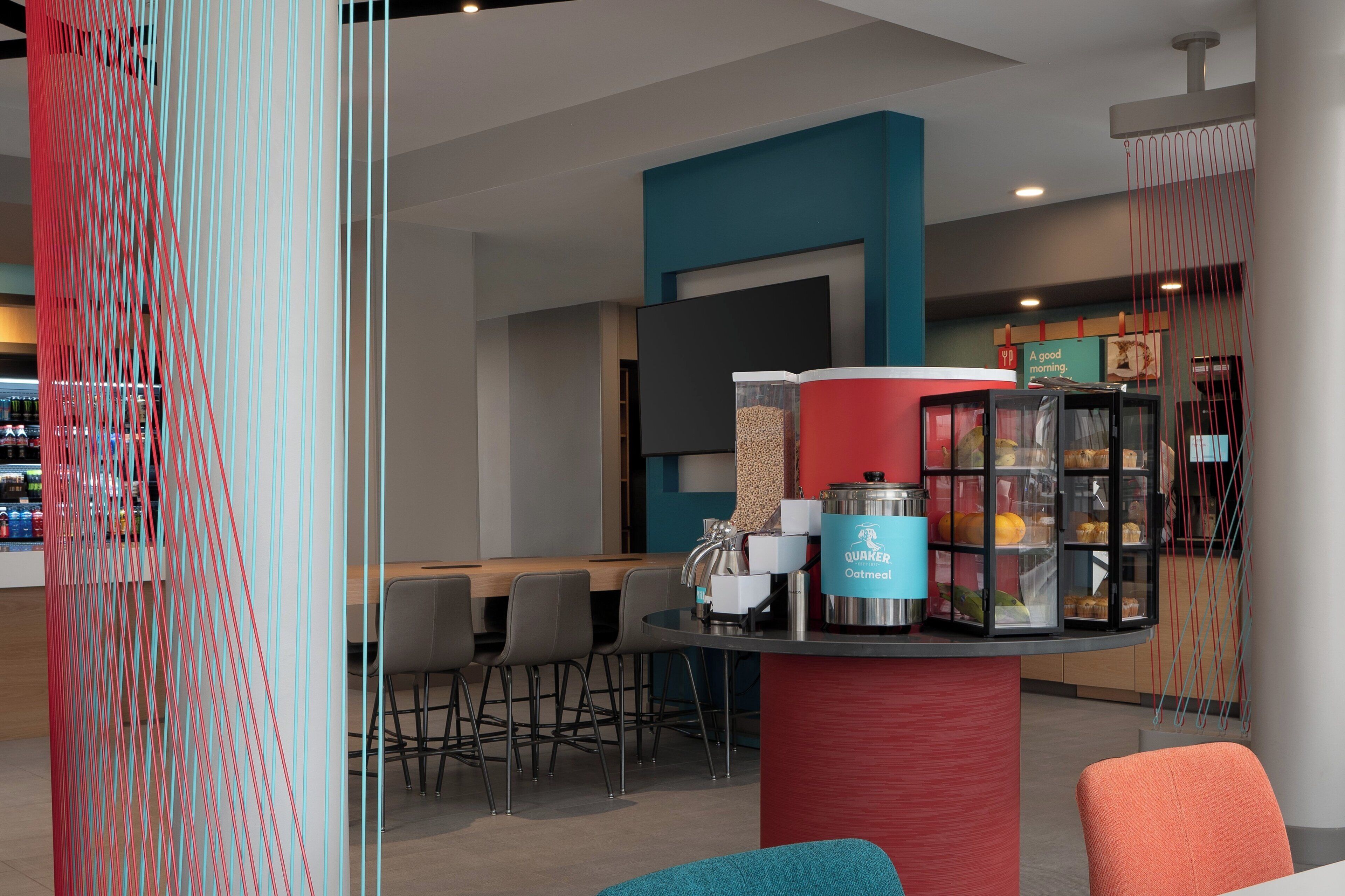 A modern cafe with a counter displaying breakfast items, a seating area with chairs, and a decorative wall with hanging elements.