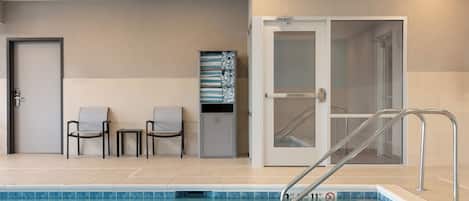 A swimming pool with a metal ladder, three chairs, a trash bin, and a door leading to a shower area.