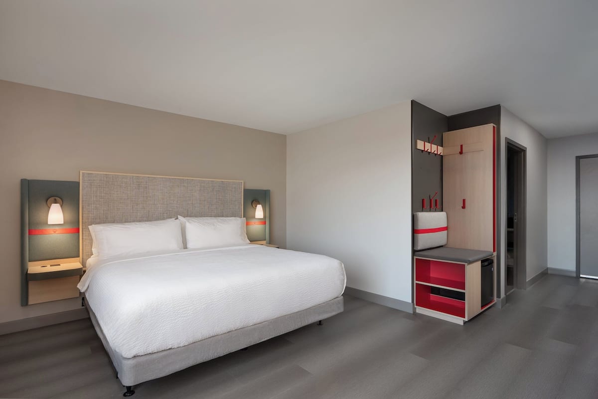 Moderni hotellihuone, jossa on suuri sänky, yöpöydät lampuineen, vaatekaappi ja sisäänrakennettu penkki.