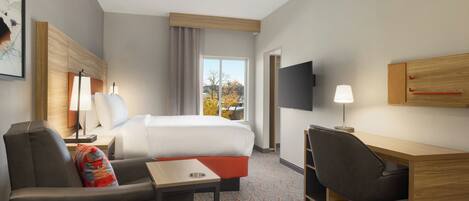 A hotel room with a bed, a desk, a chair, a TV, and a window with a view of trees.