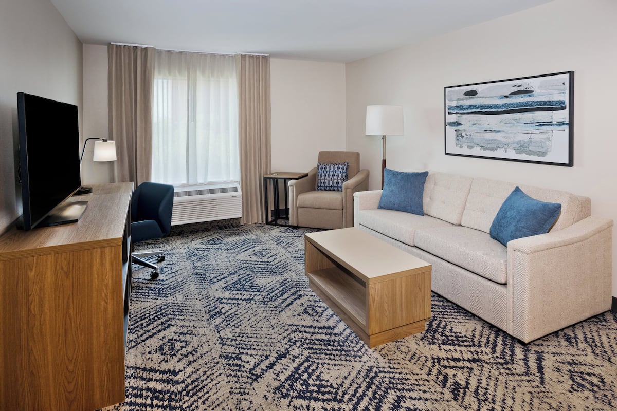 A hotel room with a wooden desk, a television, a beige sofa with blue cushions, a small side table, and a large abstract painting on the wall.