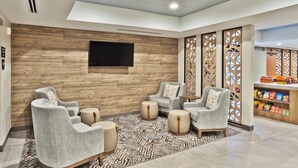 Ein moderner Lounge-Bereich mit grauen Sesseln, einem gemusterten Teppich, einem flachbildfernseher an einer Holzwand und einer Snackbar mit verschiedenen Speisen.