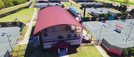 An aerial view of a sports complex with a large covered seating area, multiple tennis courts, and a parking lot.
