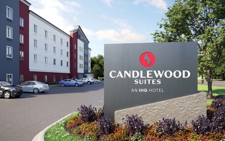 A hotel sign for Candlewood Suites, an IHG hotel, with a modern building in the background and cars parked in the lot.