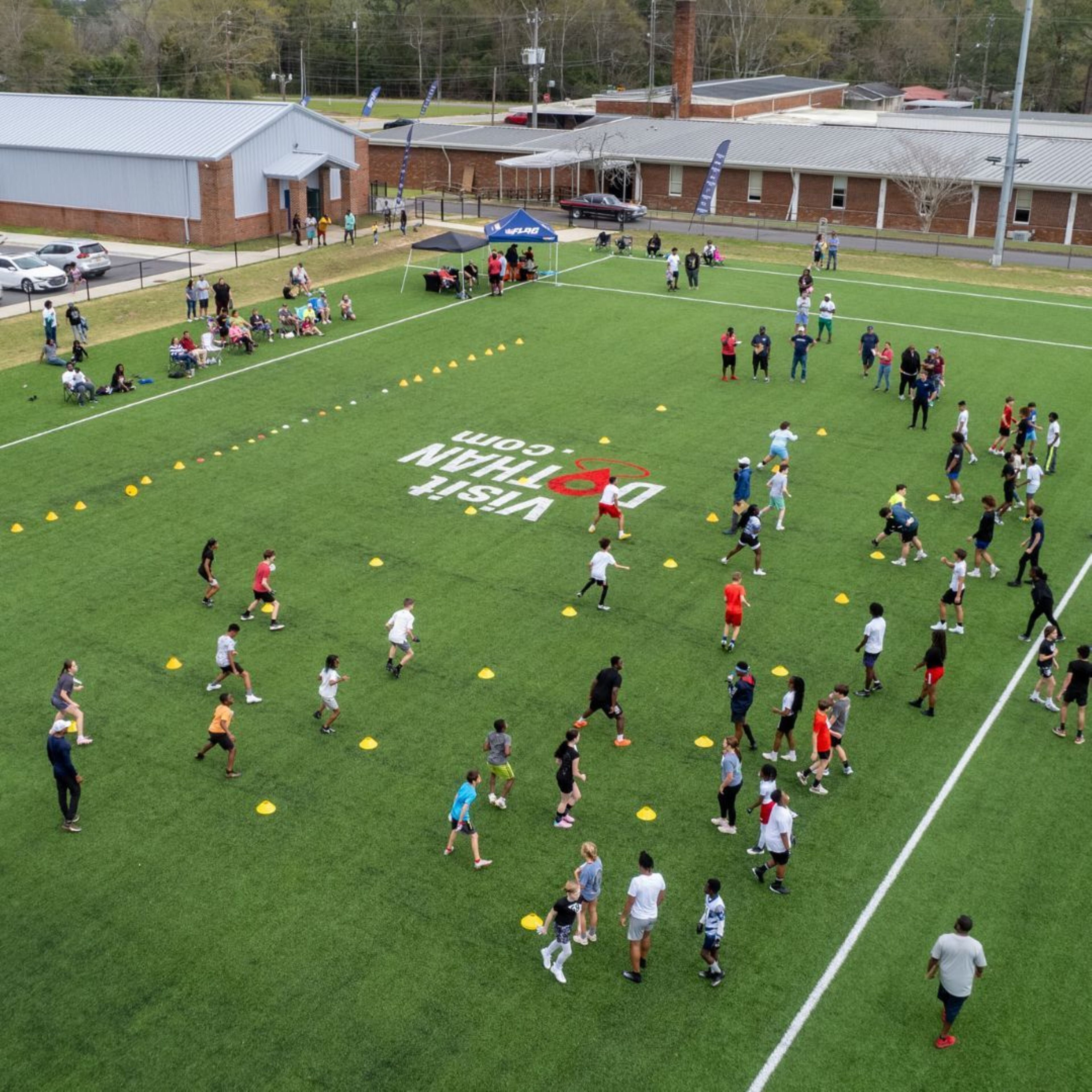 Un campo sportivo con persone che praticano diverse attività, circondato da edifici e auto parcheggiate.