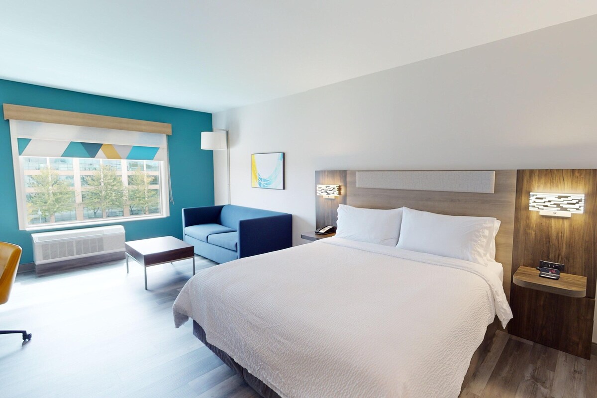 A modern hotel room with a large bed, a blue sofa, a small table, and a window with a view of trees.