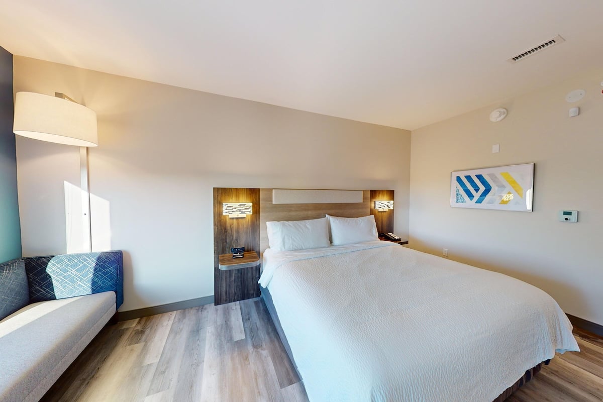 A hotel room with a large bed, a bedside table, a lamp, a chair, and a wall-mounted artwork.