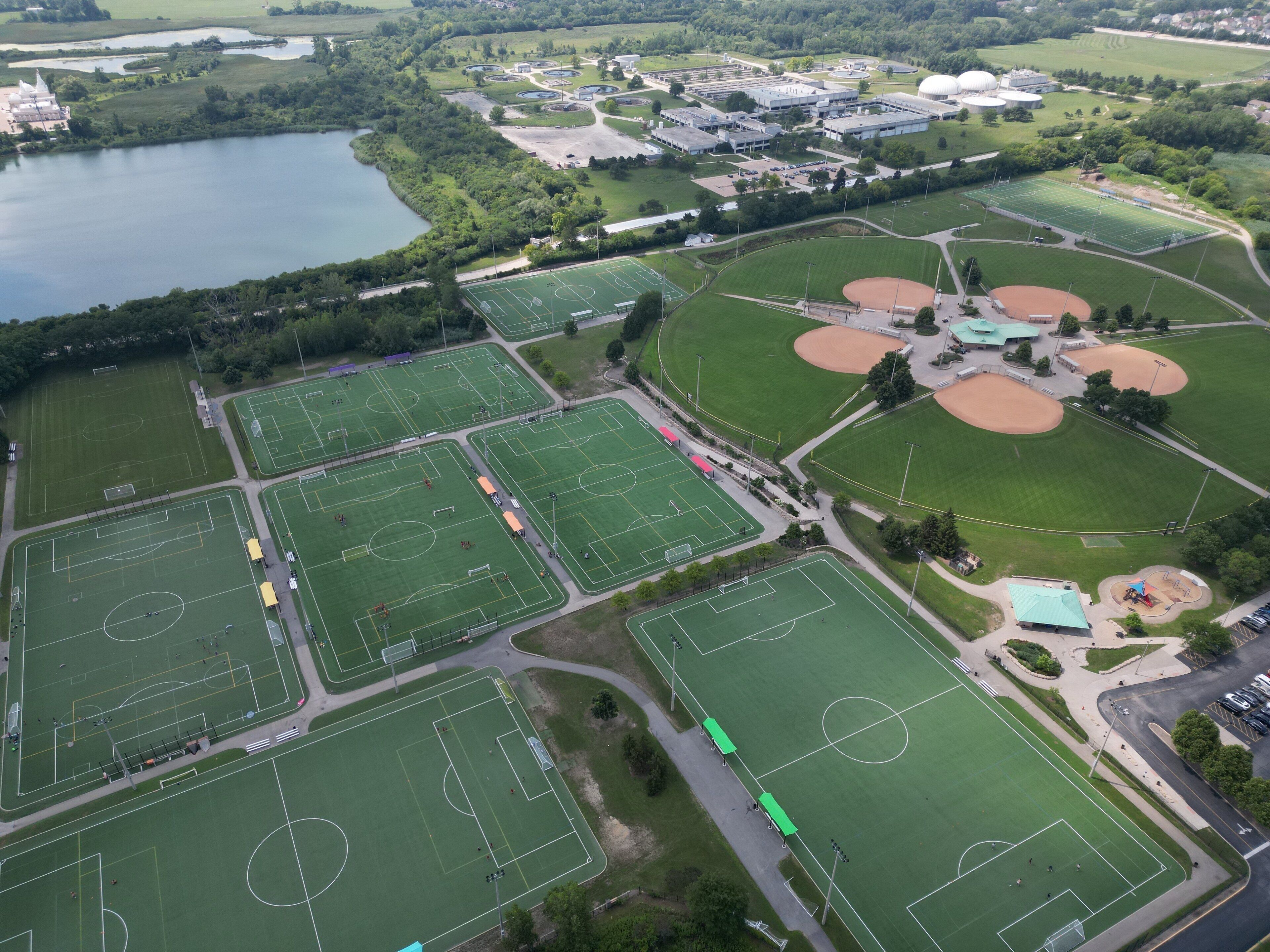 An aerial view of a sports complex with multiple fields, including soccer and baseball diamonds, surrounded by greenery and a lake.