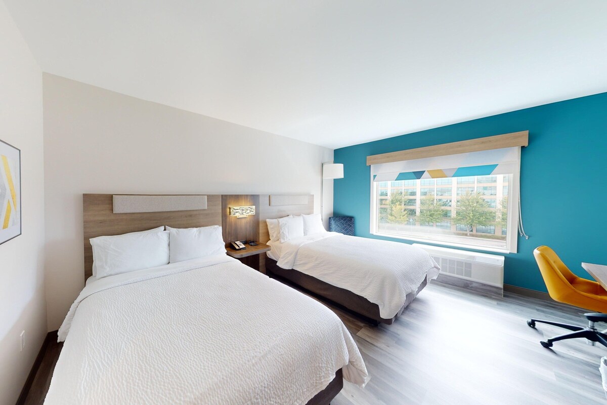 A hotel room with two beds, a large window with a view, and a yellow chair.