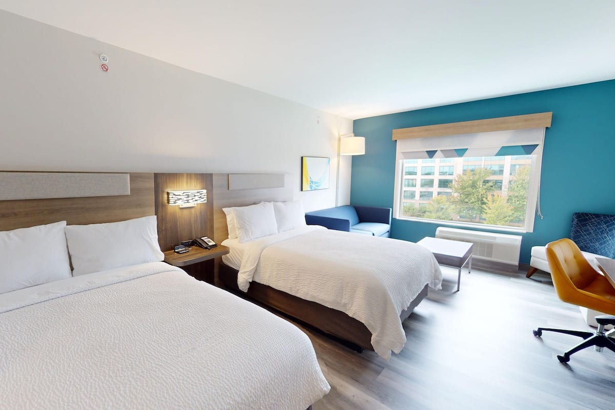 A hotel room with two beds, a desk, a chair, a window with a view, and a wall-mounted light fixture.