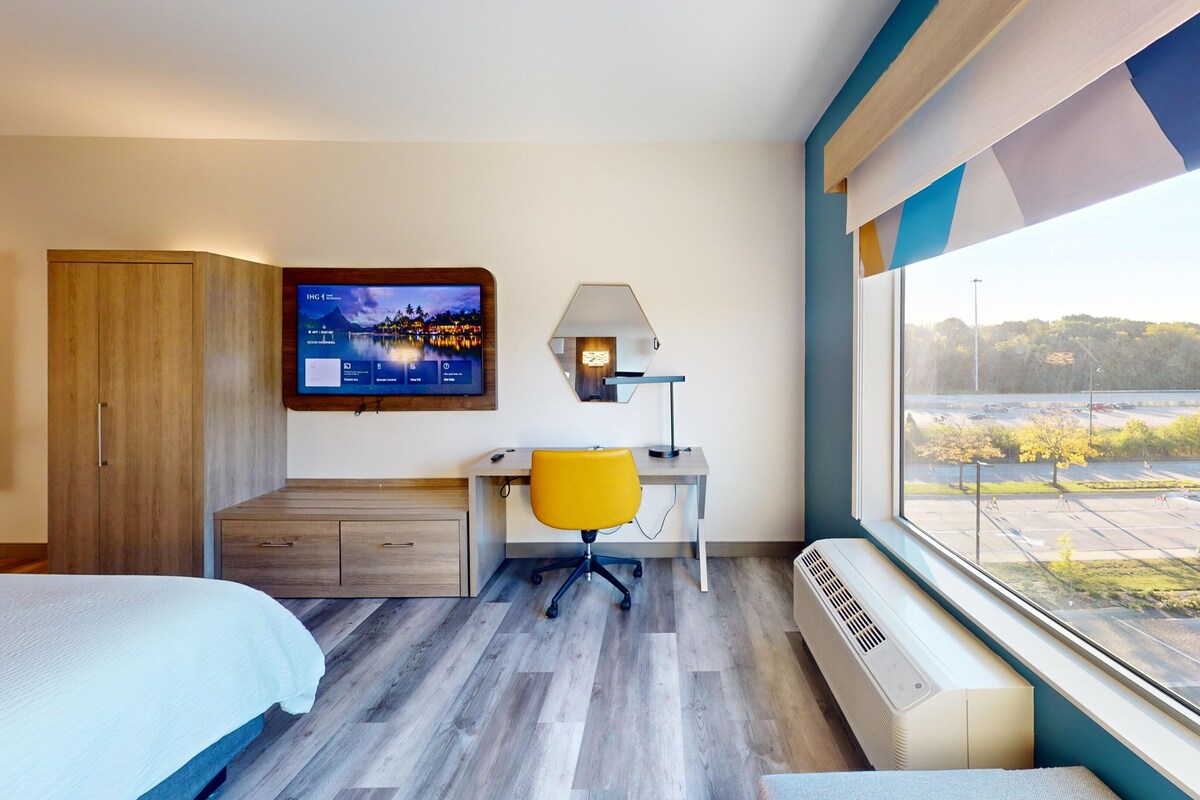 A hotel room with a bed, a desk with a chair, a TV, a lamp, and a large window with a view of a parking lot and trees.